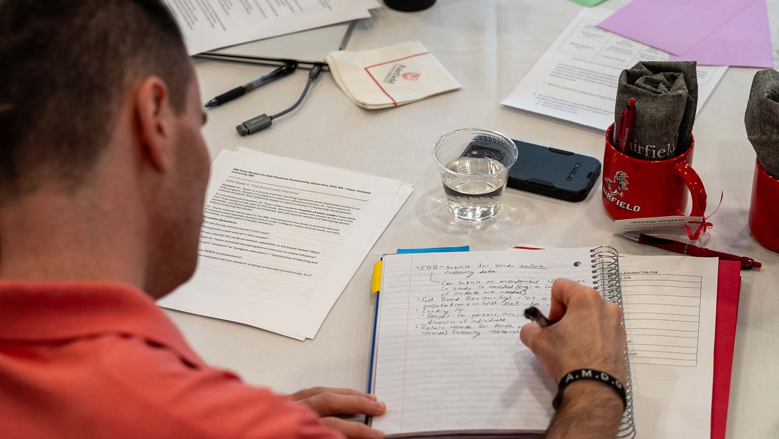 A man is focused on writing in a notebook at a table cluttered with papers and pens.