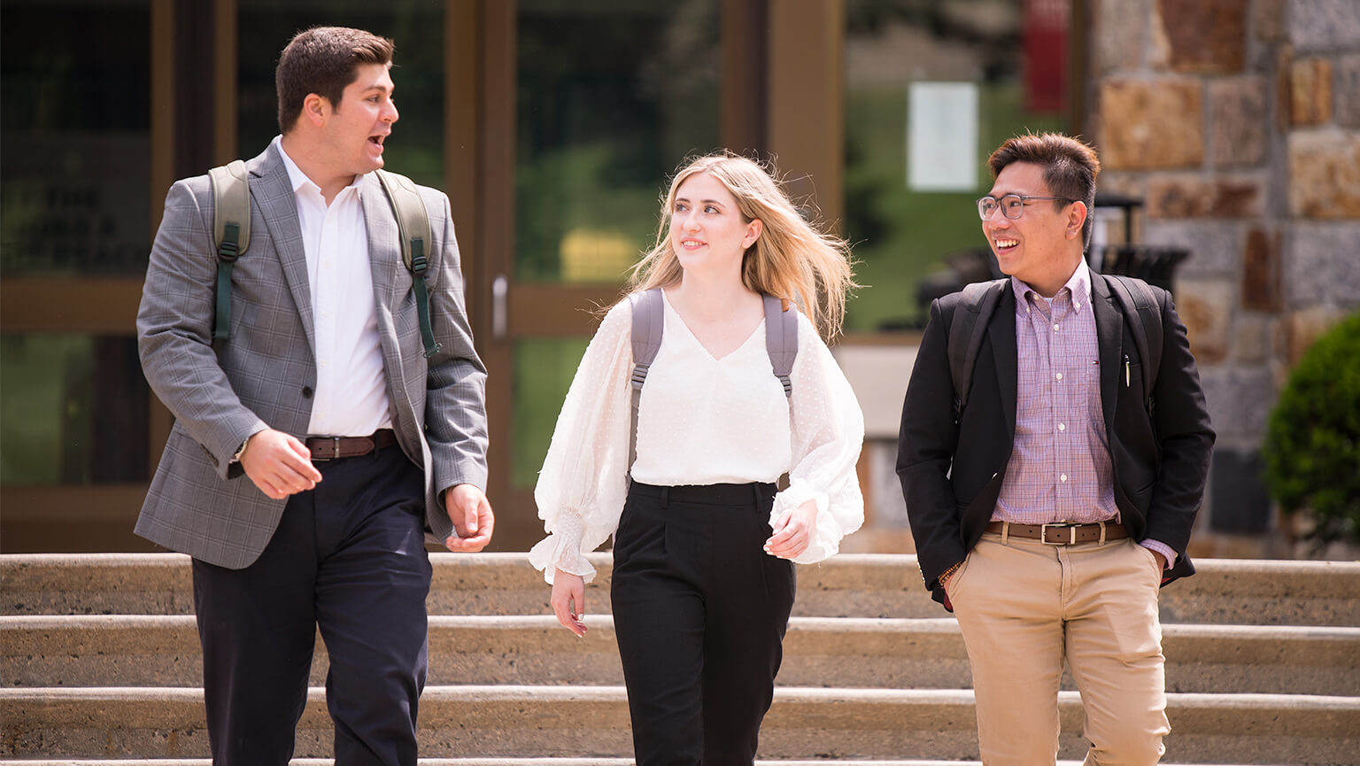 Three young adults, two men and one woman, walk together outside a building. They wear business casual attire and appear engaged in conversation, smiling.