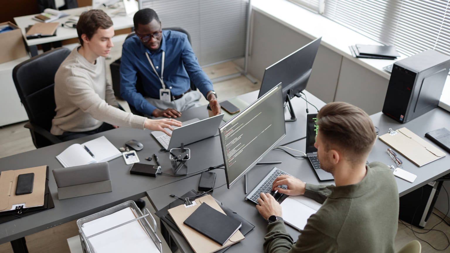 Three coworkers collaborate around computer monitors in a bright office, reviewing on-screen code and sharing ideas in a focused, friendly meeting.