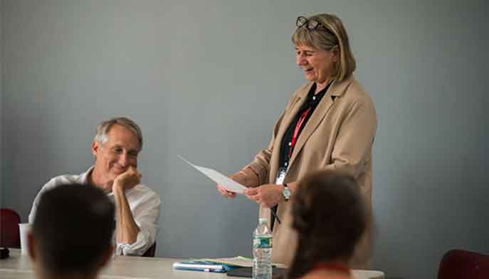 Woman standing at table speaking while people are seated listening.
