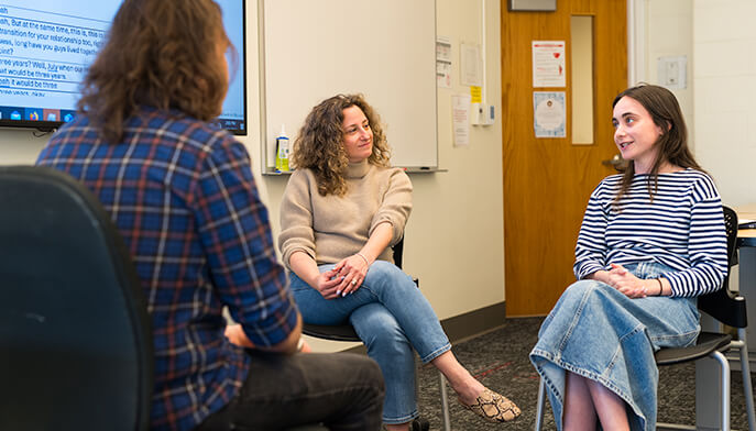 Three students engaged in conversation while seated at desks in a classroom setting.