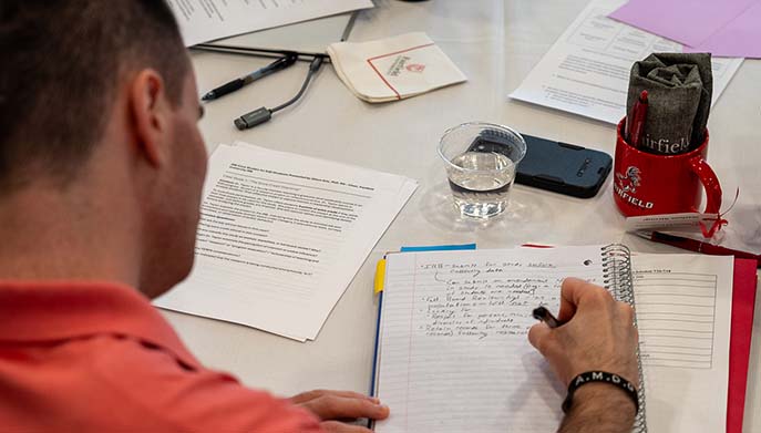 A man is focused on writing in a notebook at a table cluttered with papers and pens.
