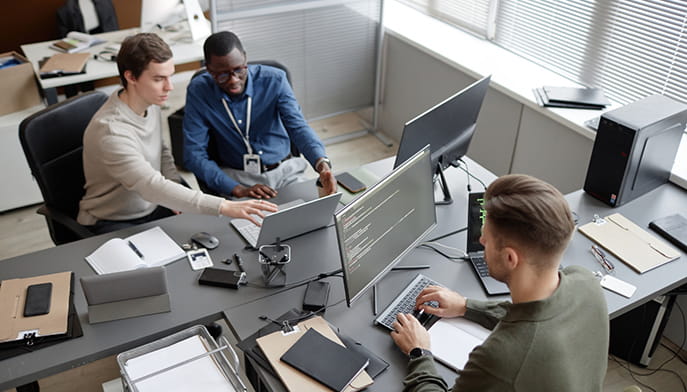 Three coworkers collaborate around computer monitors in a bright office, reviewing on-screen code and sharing ideas in a focused, friendly meeting.
