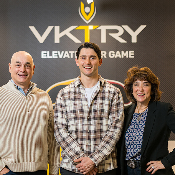 Three smiling people stand in front of a VKTRY logo with the slogan “Elevate Your Game.” The setting is professional, conveying positivity and teamwork.