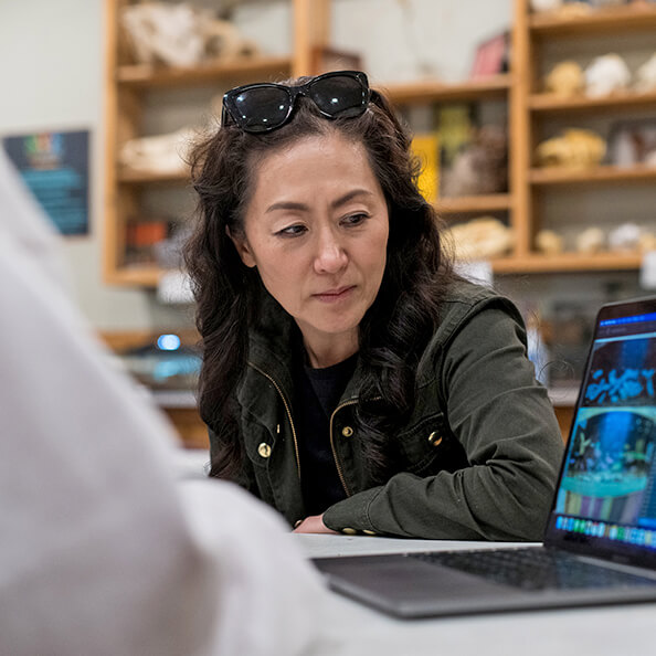 Dr. Ashley Byun’s with sunglasses on her head looks intently at a laptop screen in a classroom setting, with shelves of educational materials in the background.