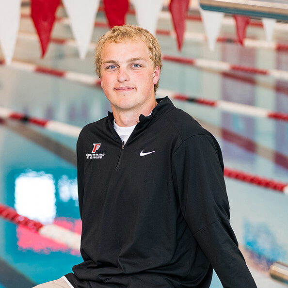 Aidan Schnapf ’26, wearing a black sports jacket sits by an indoor swimming pool with red and white lane dividers. He smiles confidently at the camera.