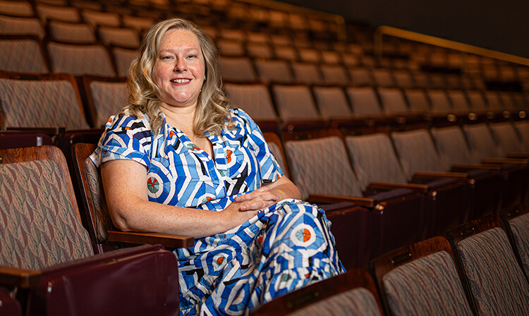 A woman with crossed arms sitting in an auditorium, appearing contemplative and engaged in the surrounding environment.