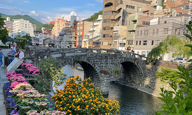 A sturdy bridge crosses a flowing river, surrounded by lush greenery and a bright sky.