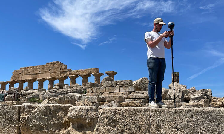 A man with a camera stands on a stone wall, surveying the view from his elevated position.