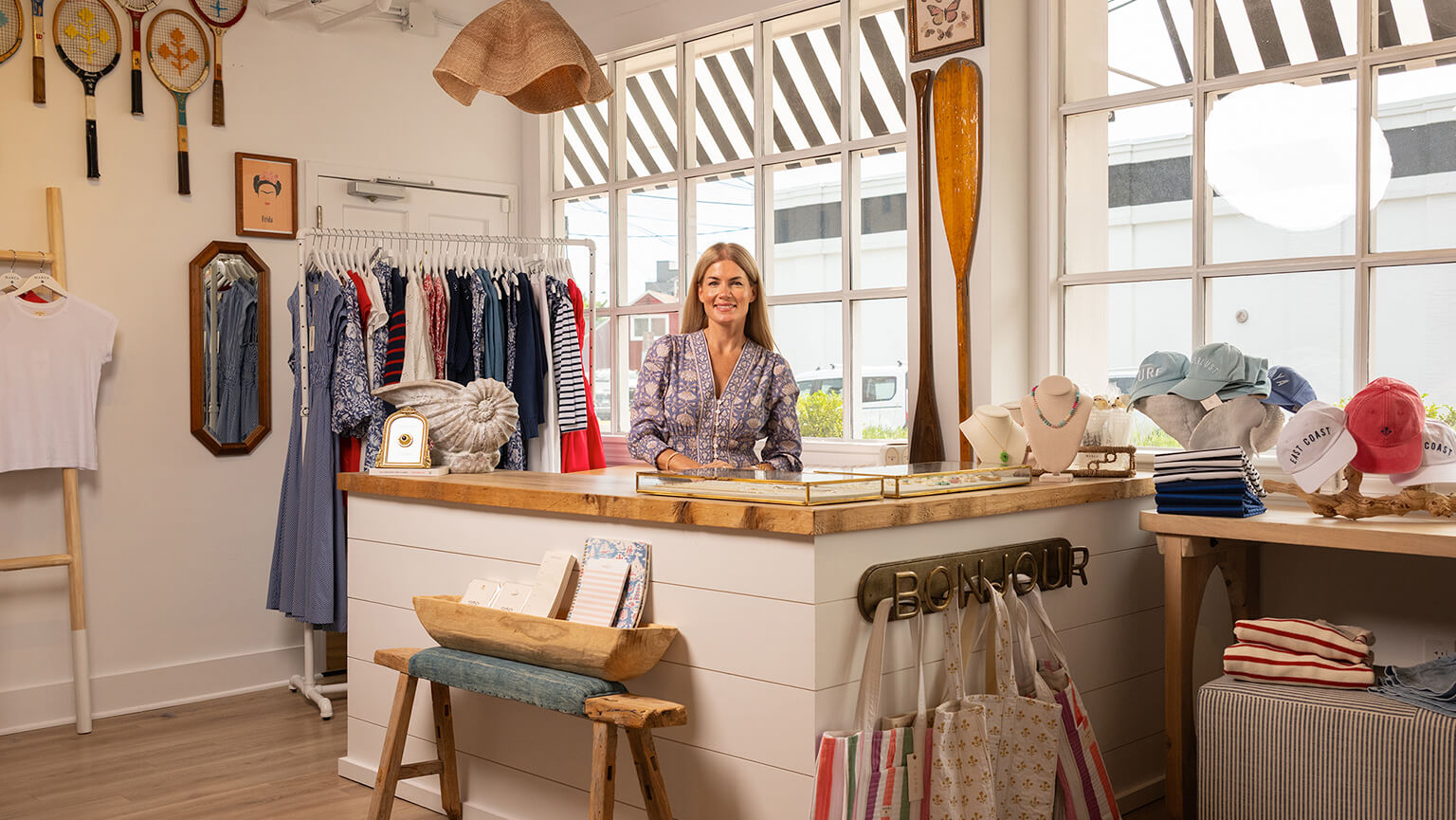 A woman stands at a counter in a clothing store, prepared to help shoppers find clothing and accessories.  