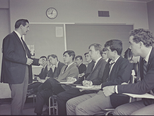 A man addresses a classroom, sharing knowledge with a group of engaged students, black and white photo.  