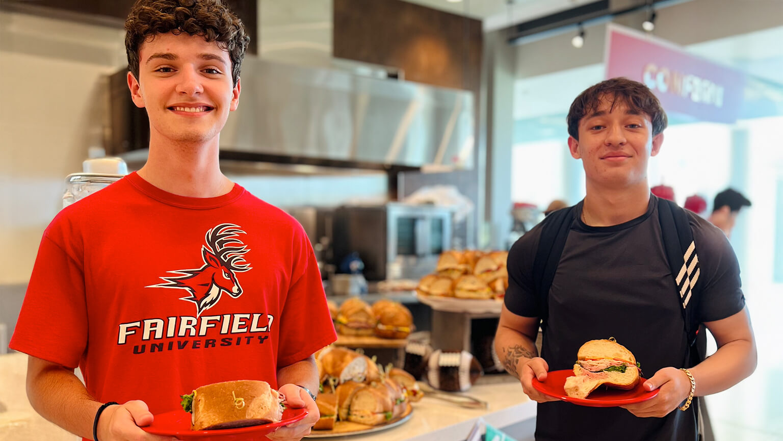 Two male students stand in front of a food station, each holding a plate filled with various foods. 