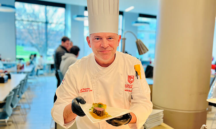 A chef in a white hat proudly holds a plate of beautifully presented food in a kitchen setting.