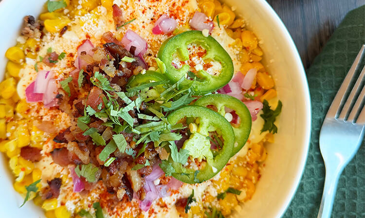 A bowl of corn topped with crispy bacon, sliced jalapenos, and melted cheese, creating a savory dish.