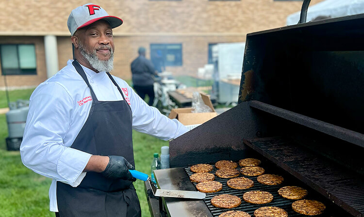 A man wearing an apron cooks burgers on a grill in an outdoor setting.
