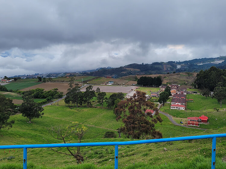 A bright blue railing frames green fields, scattered trees, and small rural houses, with distant hills beneath a cloudy sky, creating a calm, open scene.