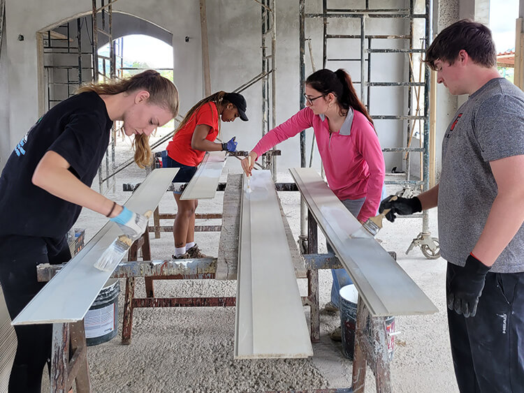 Four students in a bright workshop assemble a long metal frame, inspecting and aligning the rails; a focused, collaborative scene of hands-on construction.