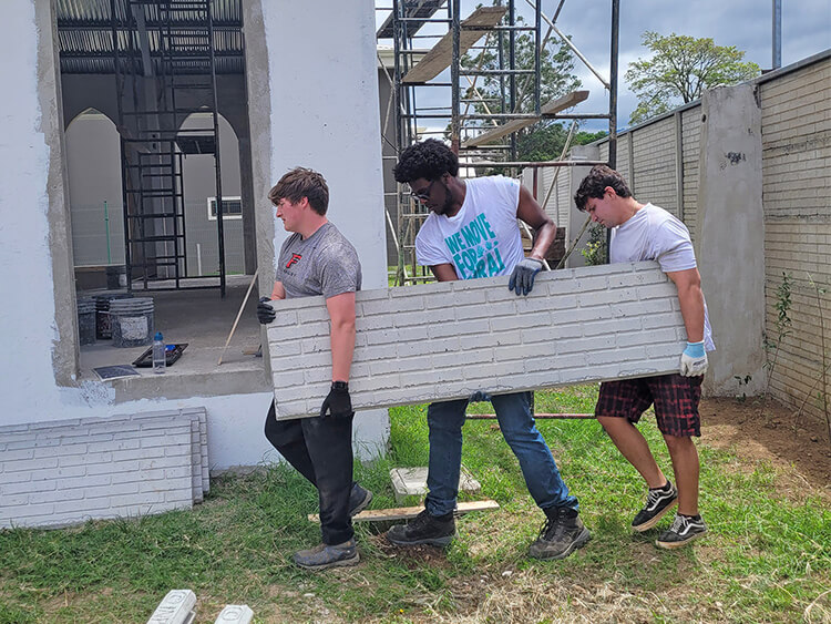 Three students lift a long gray concrete wall panel at a construction site, working together with focused expressions in an outdoor setting.