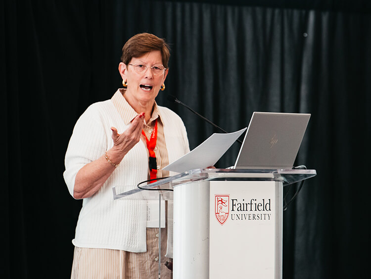 Damian Zynda, PhD in a white cardigan speaks animatedly at a Fairfield University lectern, gesturing beside an open laptop against dark curtains.