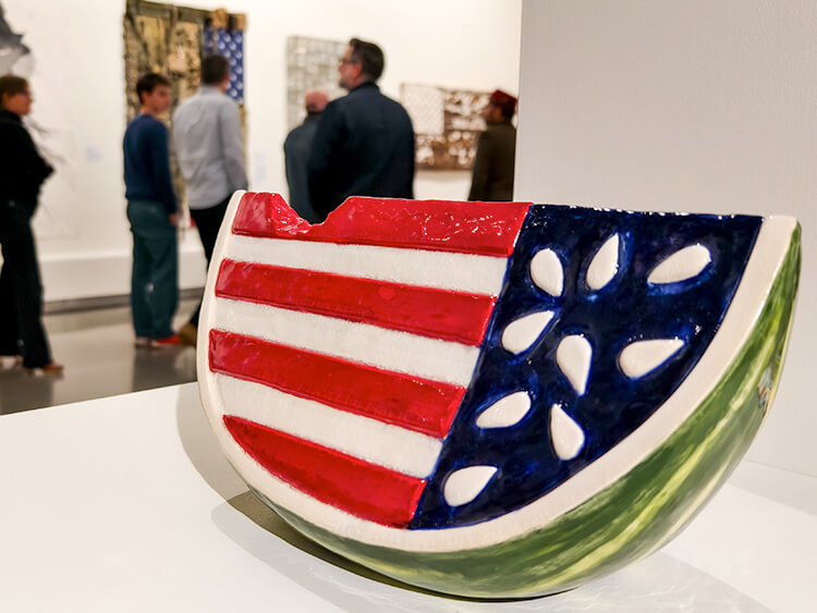 A sculpture resembling a watermelon slice painted with a U.S. flag design is in the foreground. In the background, people observe art in a gallery.