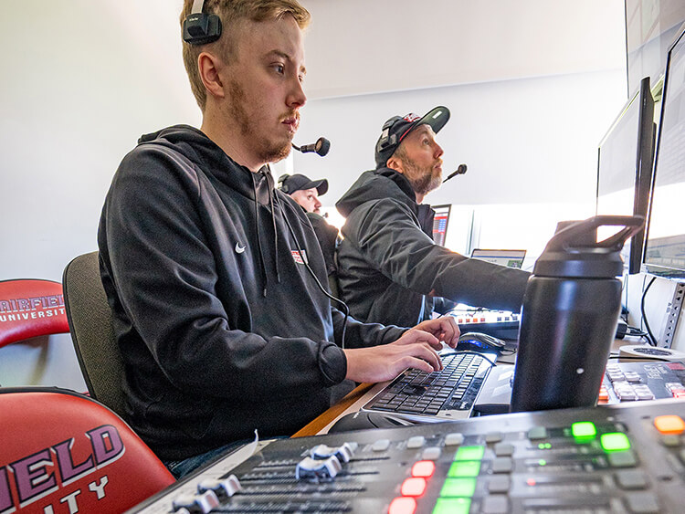 Two men wearing headsets sit at a control desk, focused on computer screens. The atmosphere is professional and attentive, with equipment nearby.
