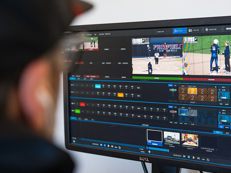 A person views a computer screen displaying video editing software. The screen shows multiple video thumbnails of a softball game. The mood is focused.