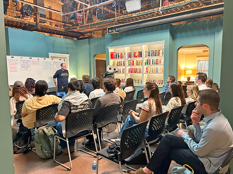 Classroom scene with a lecturer in front of students seated on chairs. A whiteboard with notes is visible, alongside bookshelves and a camera setup. The atmosphere is focused.