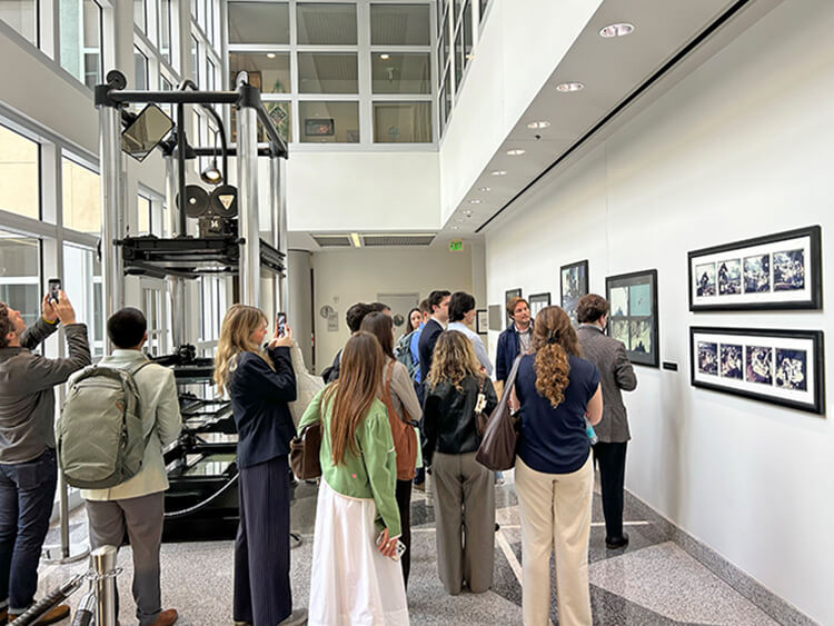 A group of students in an art gallery observe framed photographs on a white wall. Some use smartphones for photos. The setting is bright and modern.