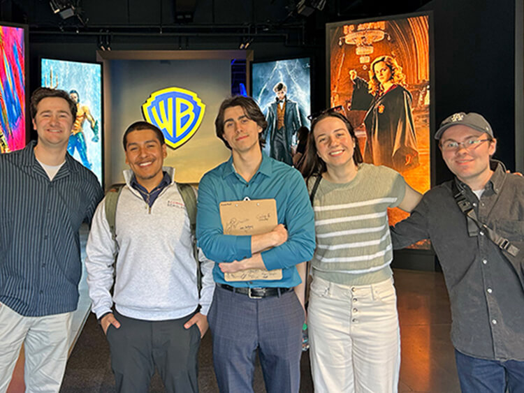 A group of five students smiling in front of a Warner Bros. logo. They are standing indoors, flanked by colorful movie posters, creating a lively and enthusiastic atmosphere.