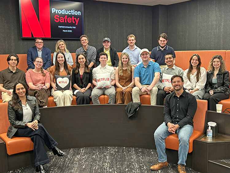 A group of 17 people, casually dressed, sit in a tiered seating area. Two hold Netflix logos. A screen displays "Production Safety" and "Fairfield University Visit" The setting is modern and professional, with a positive, collaborative atmosphere.