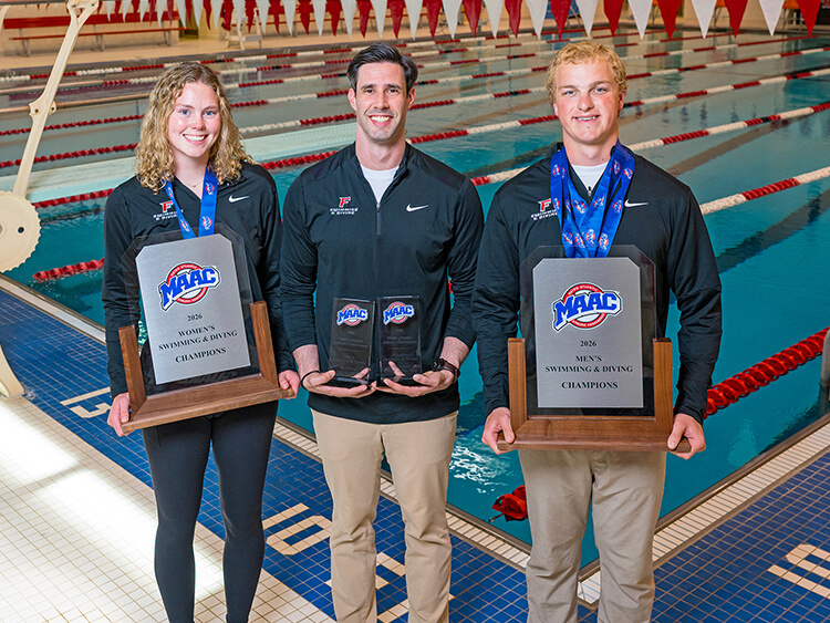 Three smiling individuals in black athletic wear stand poolside holding MAAC swimming and diving championship trophies. The background is a brightly lit indoor pool.