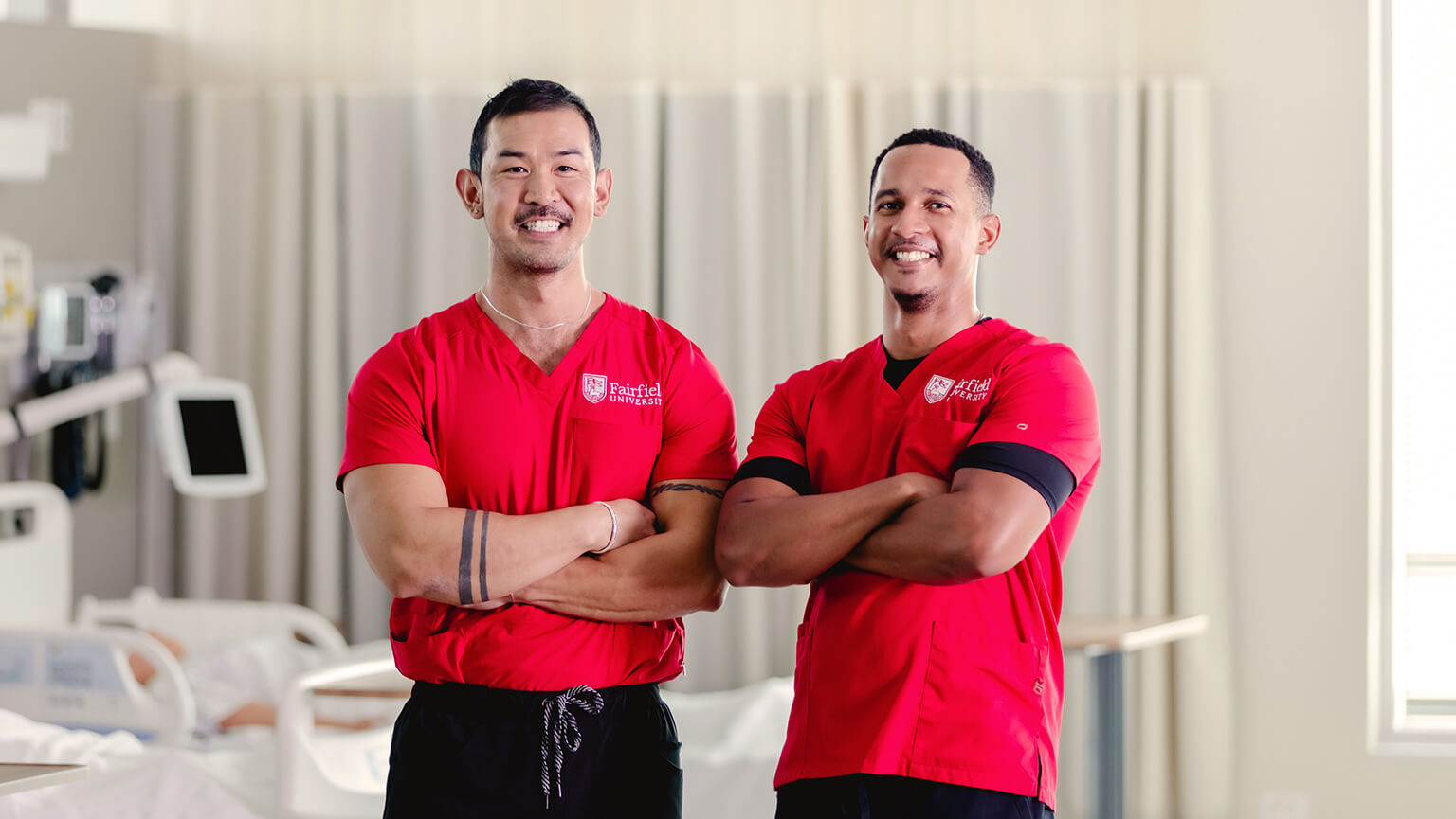 Two male nursing professionals in red scrubs stand confidently with arms crossed in a hospital setting.
