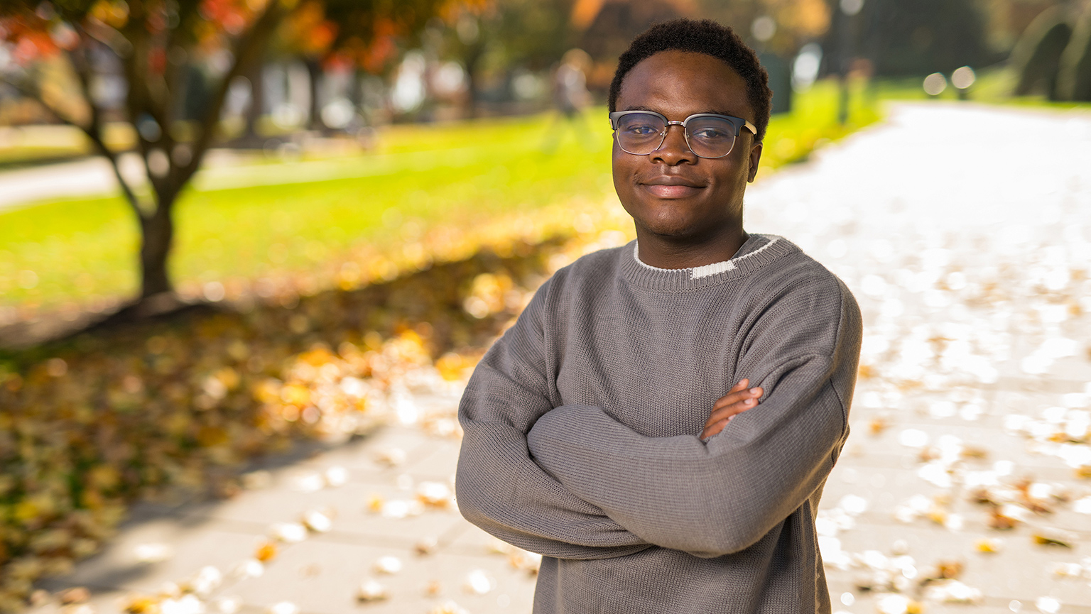 A young man with glasses poses on Fairfield University campus, showcasing a serene environment filled with lush trees and grass.