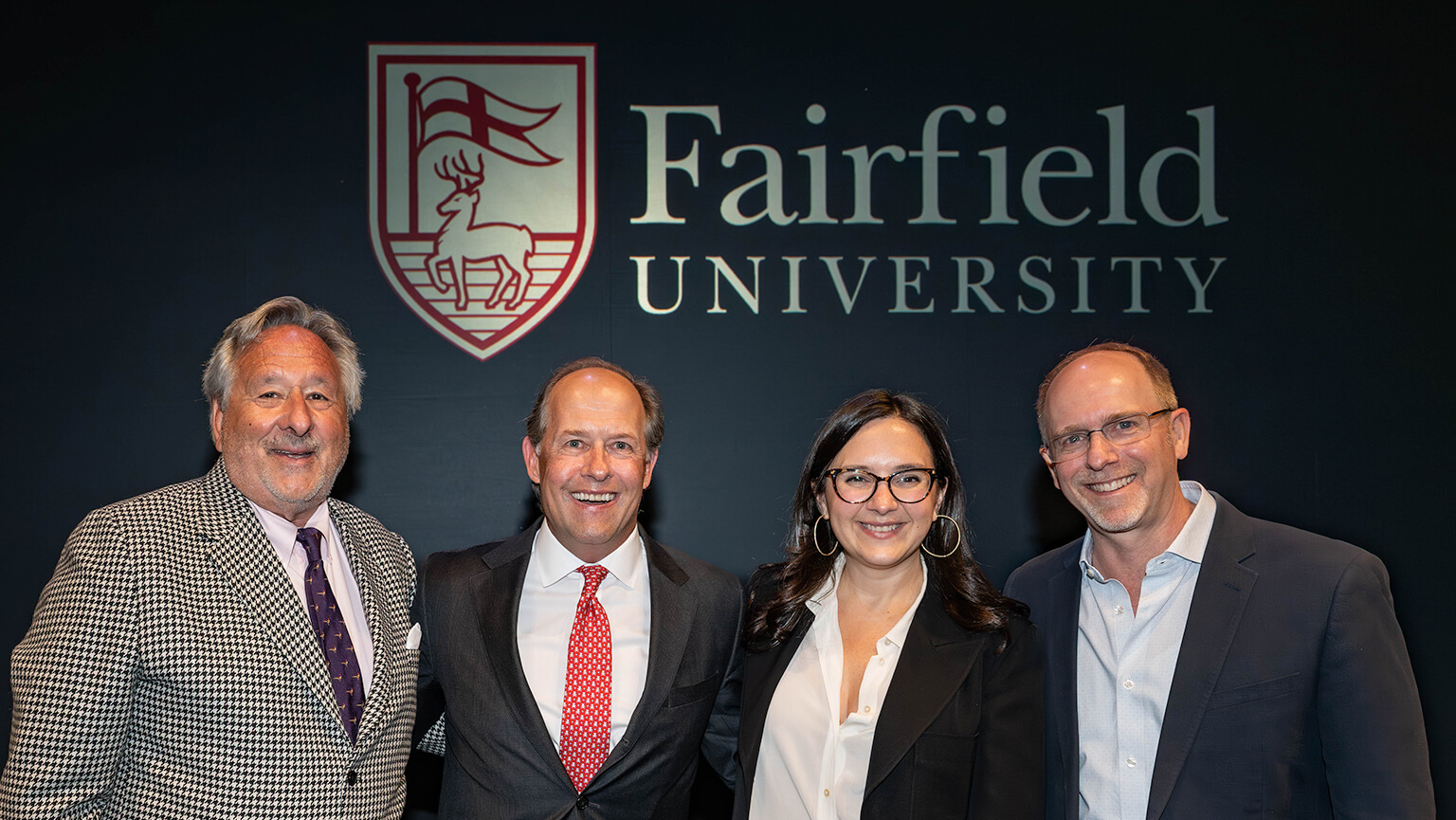 Four people are gathered in front of the Fairfield University logo, highlighting their connection to the institution.