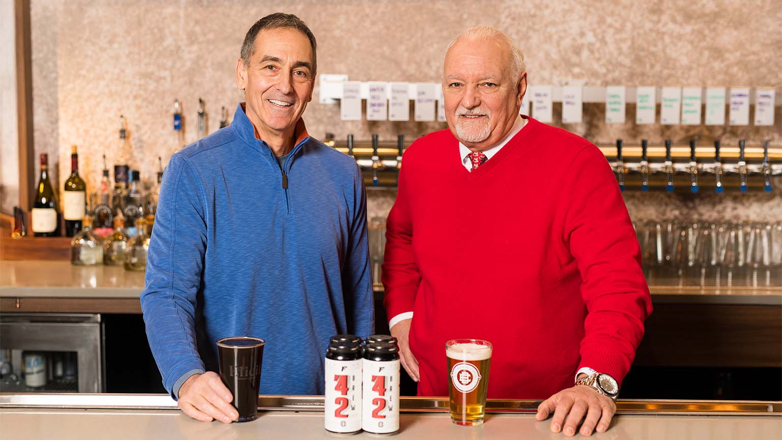 Image of Two friendly men at a bar, standing behind a counter filled with various beers and drinks.