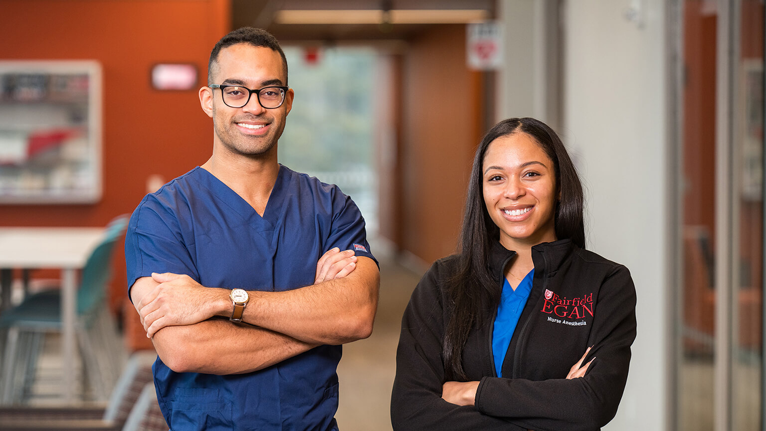 Two individuals wearing scrubs stand in a brightly lit office hallway, discussing something with focused expressions.