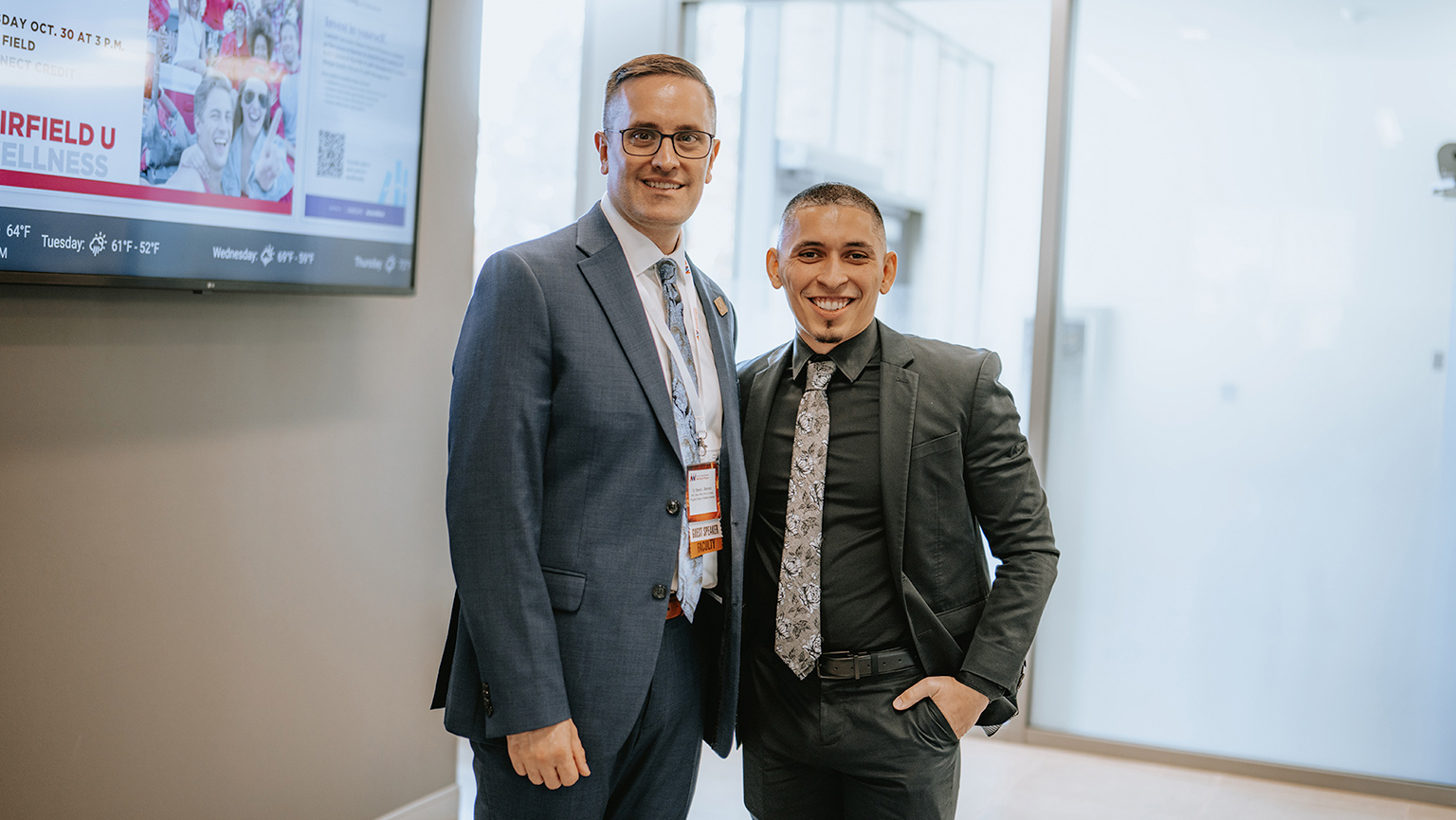 Two men in suits standing side by side, both smiling and looking towards the camera. 