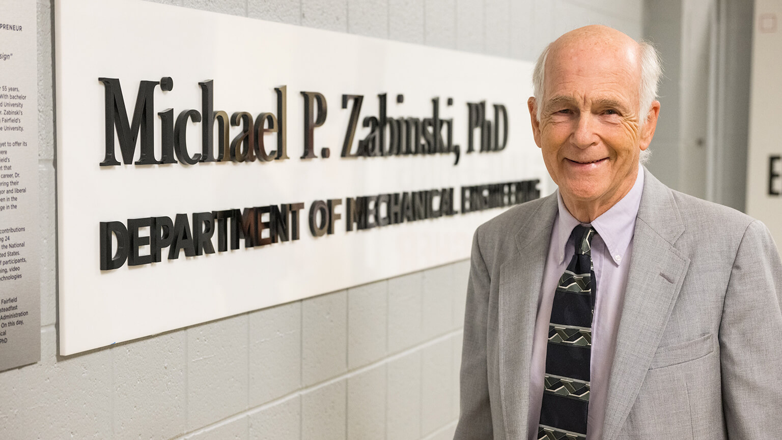 Portrait of Michael P. Zabinski, PhD standing next to a sign with his name on it.
