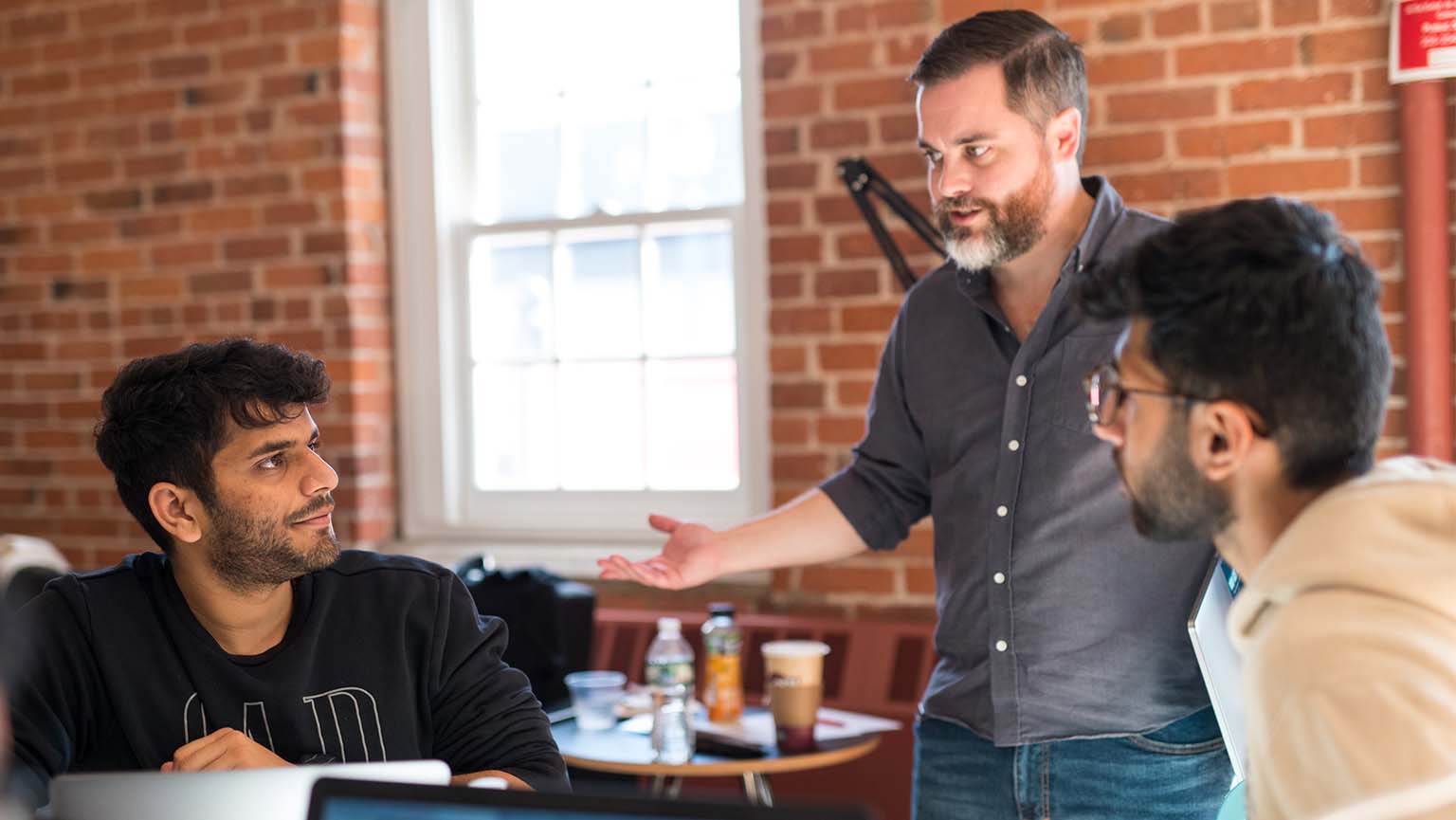 Image of three men sit around a table, each focused on their laptops, engaged in a collaborative discussion.
