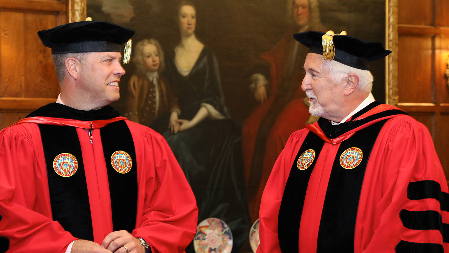 Two men wearing red robes and black hats stand side by side, showcasing a traditional or ceremonial attire.