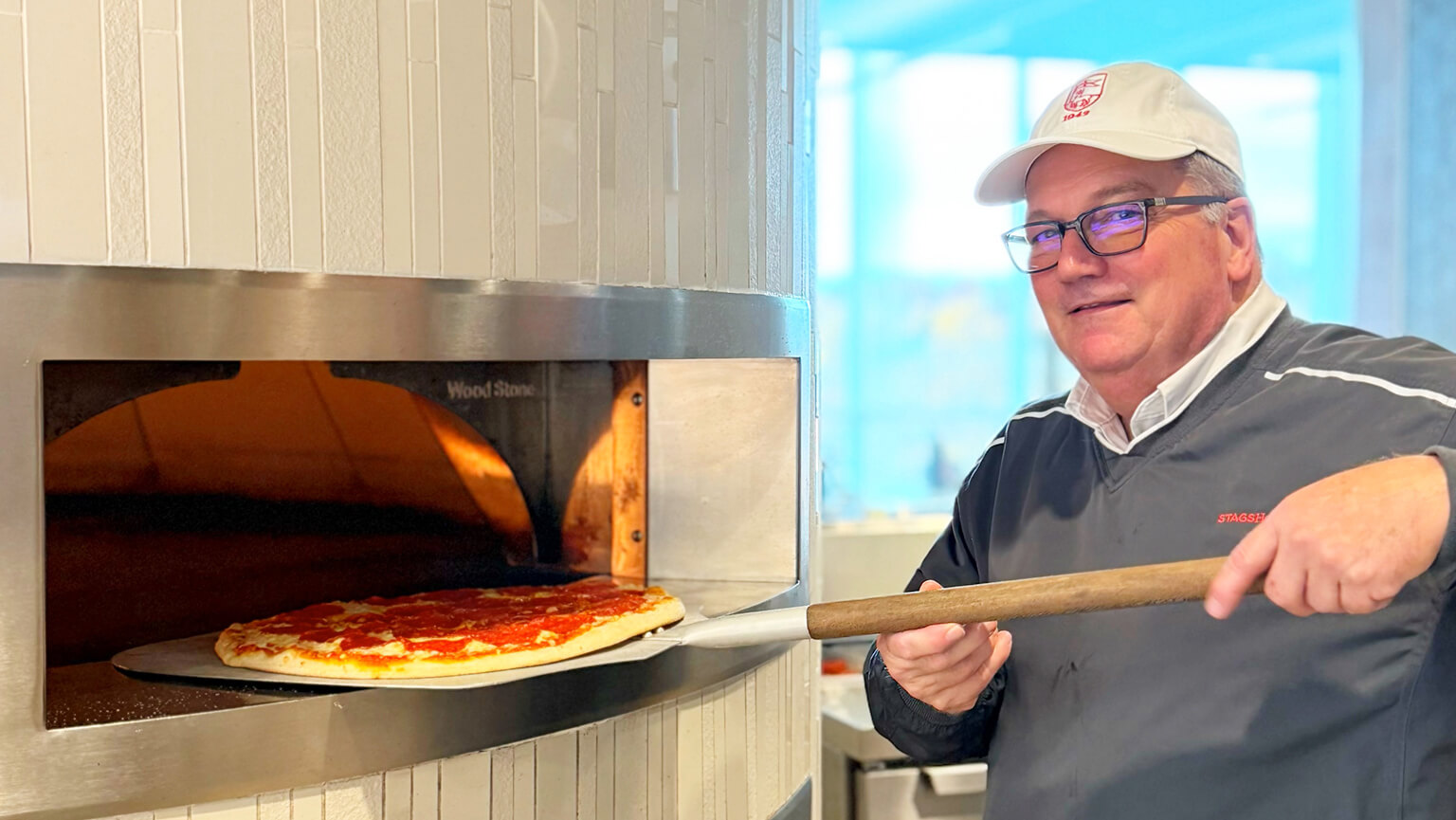 Jay DeGioia wearing a gray shirt and glasses placing a pizza into a pizza oven. 