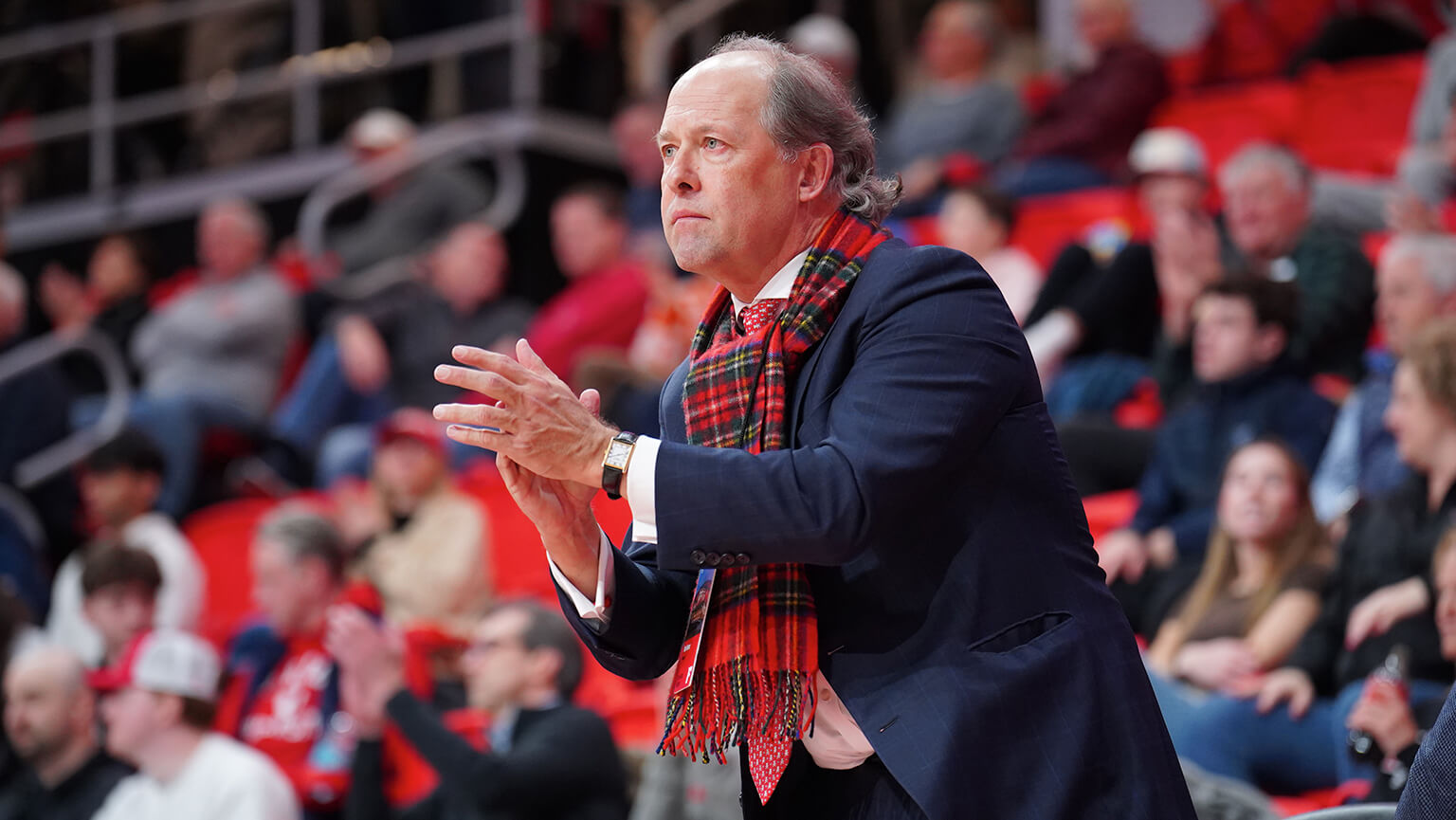 President Mark Nemec in a suit and tie stands on the sidelines, observing the basketball game with a focused expression.