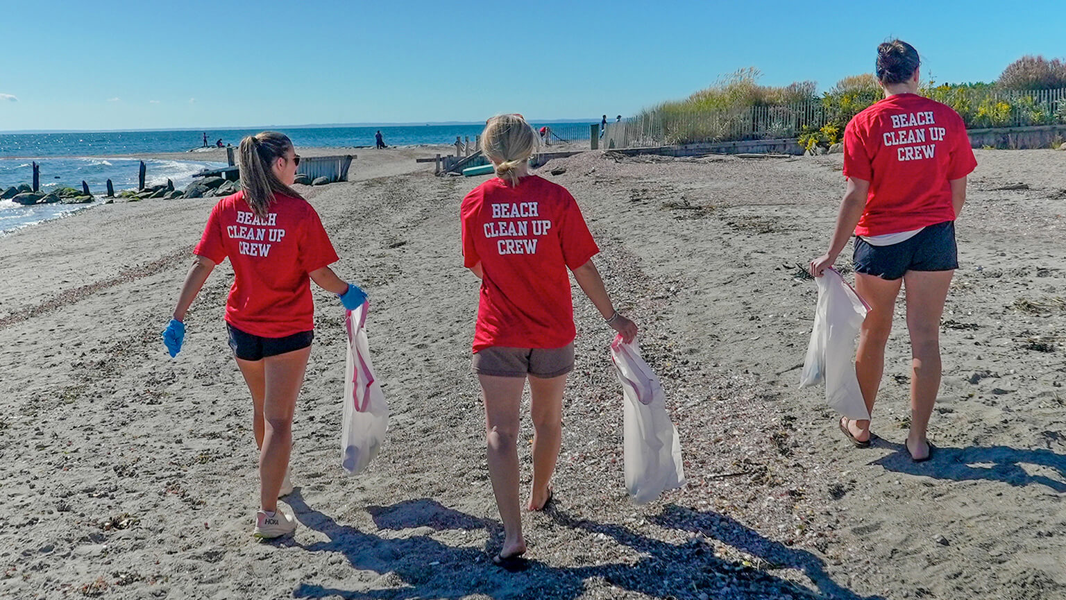 Three female students in red shirts walk along the beach, collecting trash in bags to clean up the shoreline.  