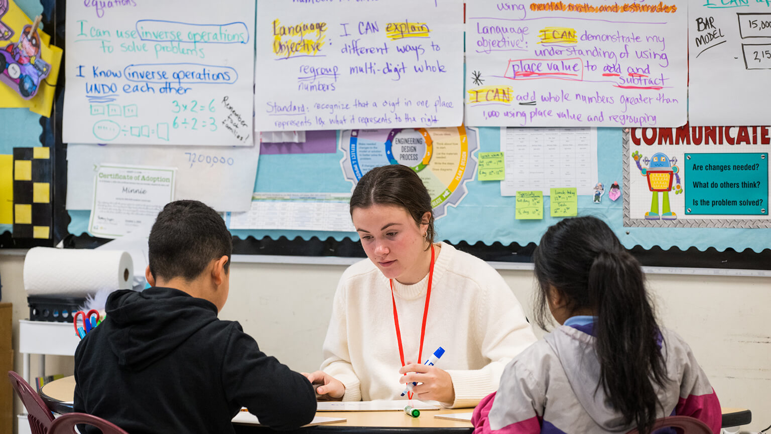 A student teacher assists students with their work in a bright, engaging classroom setting filled with educational materials.