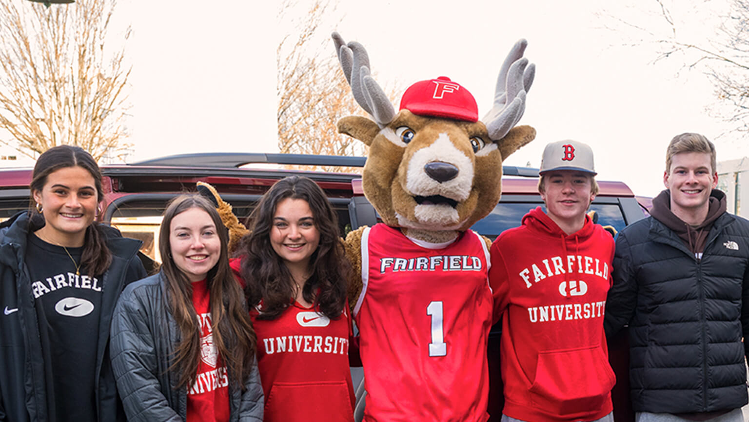 Several students stand together, smiling with a cheerful Lucas the Stag mascot at a community gathering.  