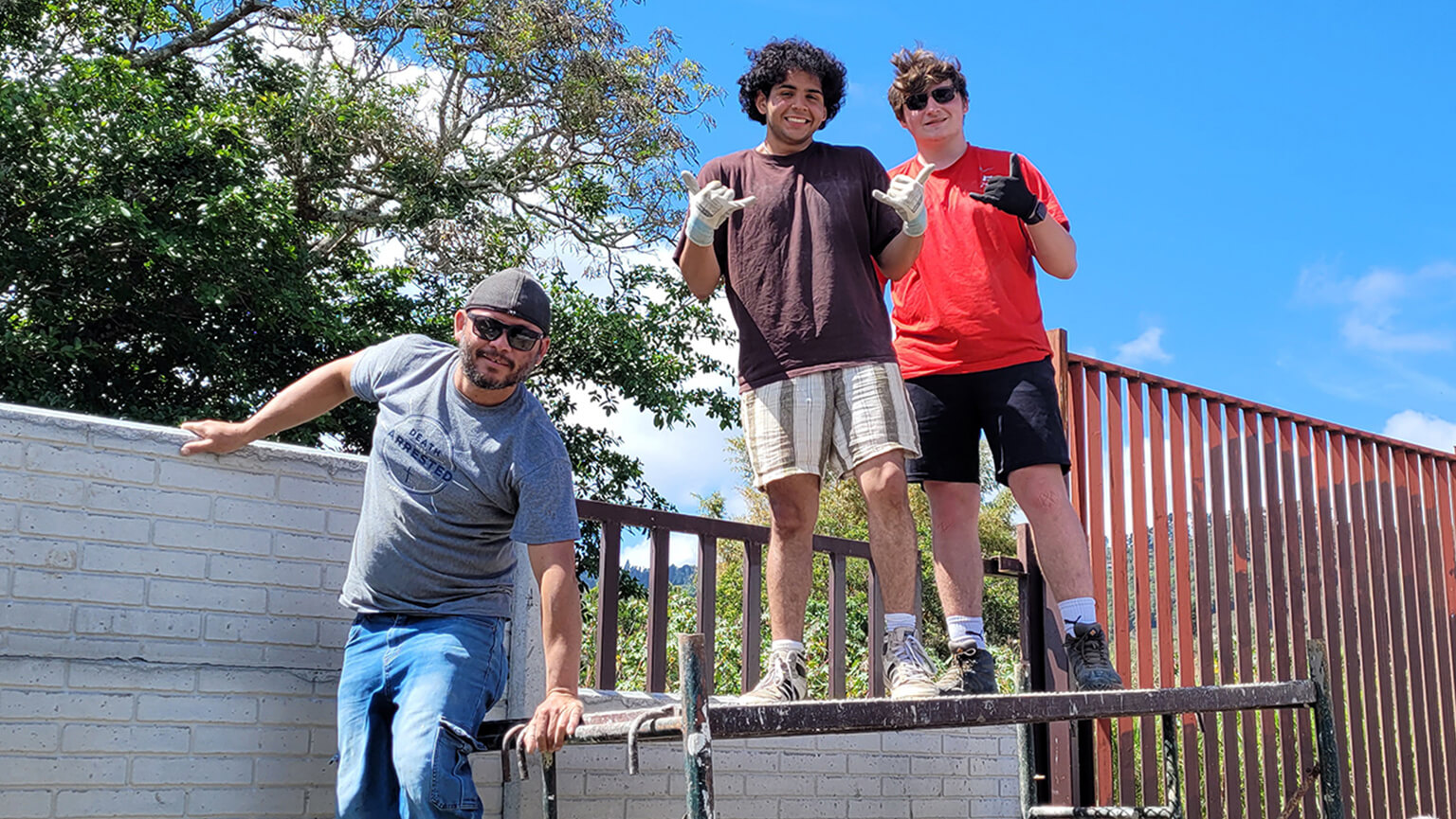Three students pose on a raised platform by a railing, smiling in casual streetwear under a bright blue sky; the scene feels upbeat and relaxed.