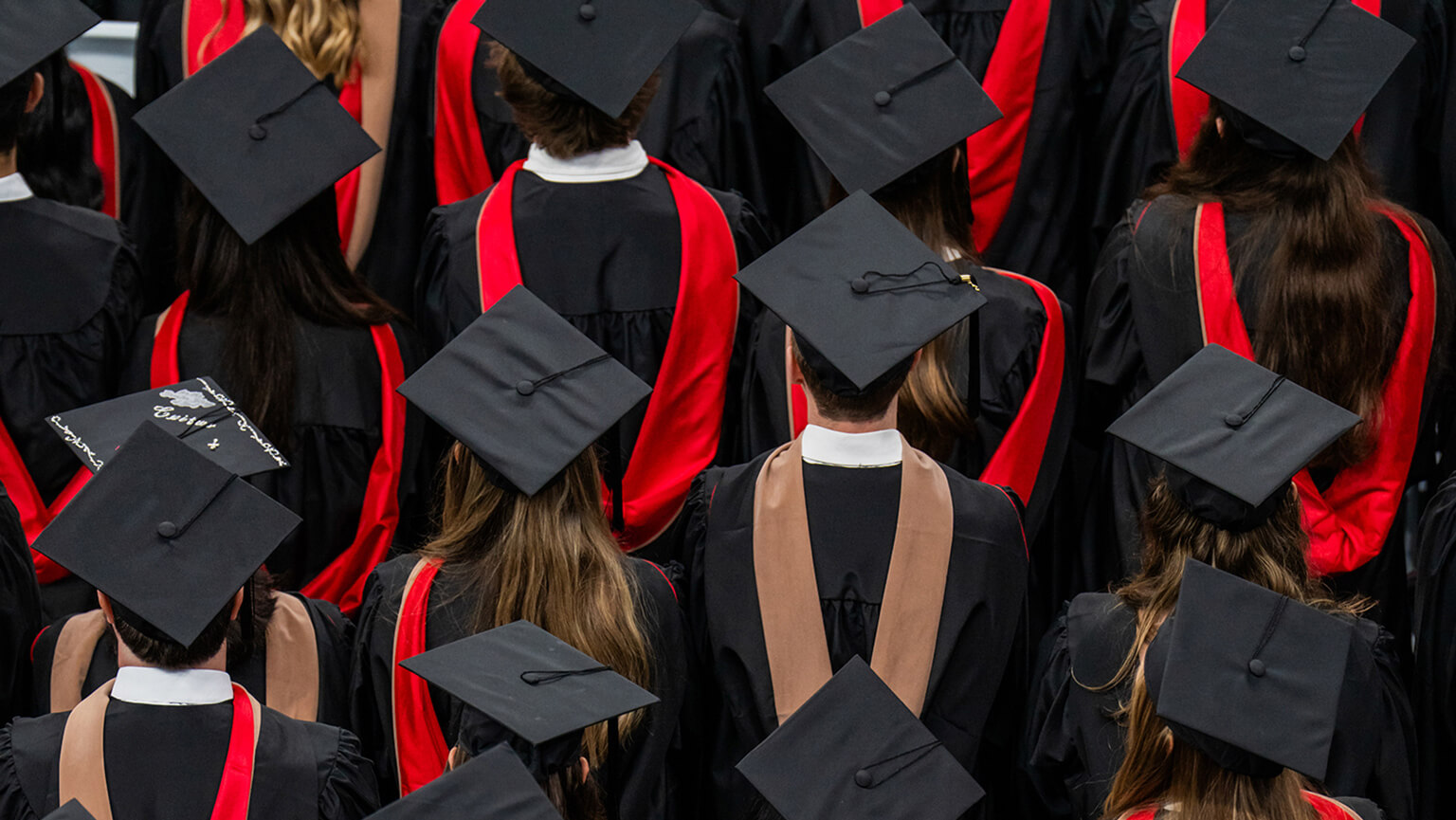Overhead view of graduates in black caps and gowns with red accents, gathered closely at a graduation ceremony in a celebratory mood.