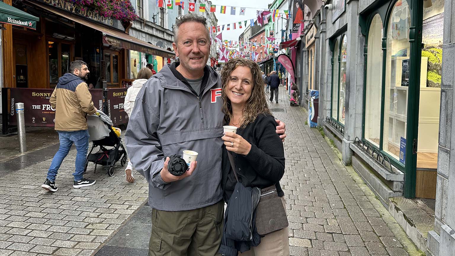 Donald and Karen Wilson P’26 A stand arm in arm on a lively cobbled shopping street, holding a camera and takeaway cups beneath colorful flags.