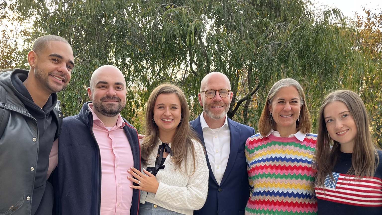 Six smiling adults pose together outdoors beneath leafy trees, dressed in casual and smart clothing, including a bright striped sweater and flag-print top; warm, friendly group portrait.
