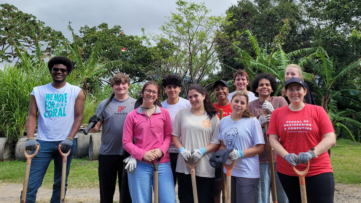 A smiling group of students stand outdoors with shovels, surrounded by trees and greenery, ready for a community gardening project.